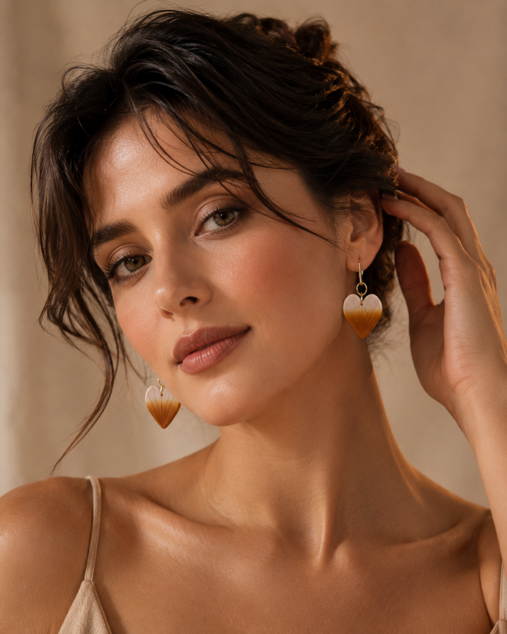 A close-up lifestyle shot of a woman wearing heart-shaped dangle earrings with a gradient design transitioning from white to warm amber-brown, set against a soft beige background.