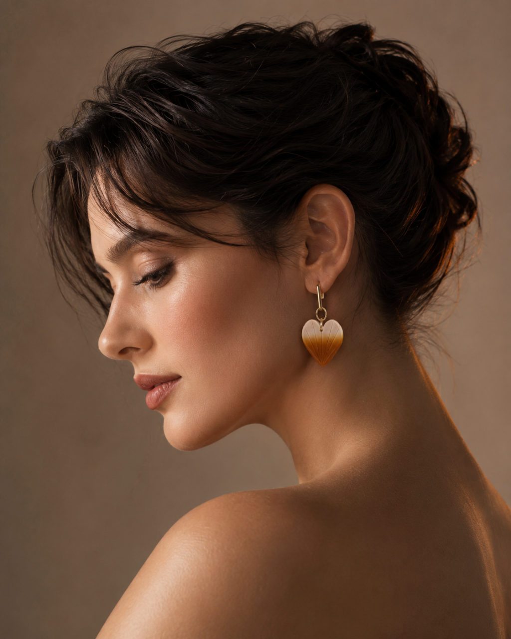 A woman in profile wearing a dangling heart-shaped gold earring with a cream-colored center, set against a warm brown studio background.