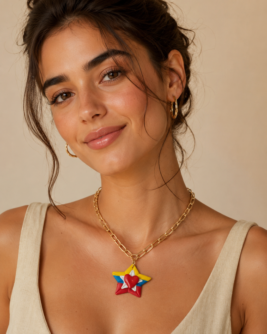 A woman wearing a gold chain necklace with a colorful star-shaped pendant in red, yellow, and blue, paired with gold hoop earrings and a beige top against a warm beige background.