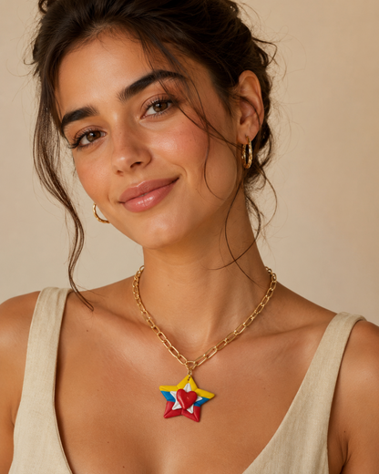 A woman wearing a gold chain necklace with a colorful star-shaped pendant in red, yellow, and blue, paired with gold hoop earrings and a beige top against a warm beige background.